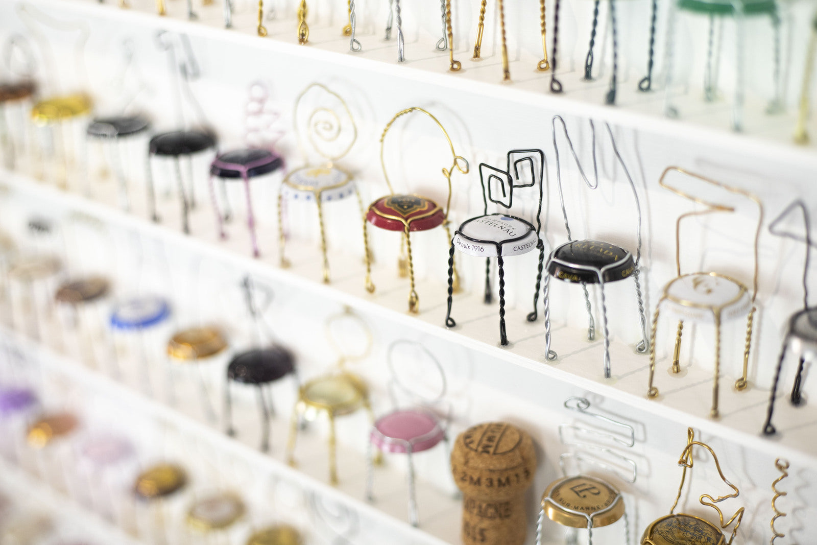 Close-up of Joanne Tinker's miniature chair sculptures made from recycled champagne bottle tops, displayed on white shelves.