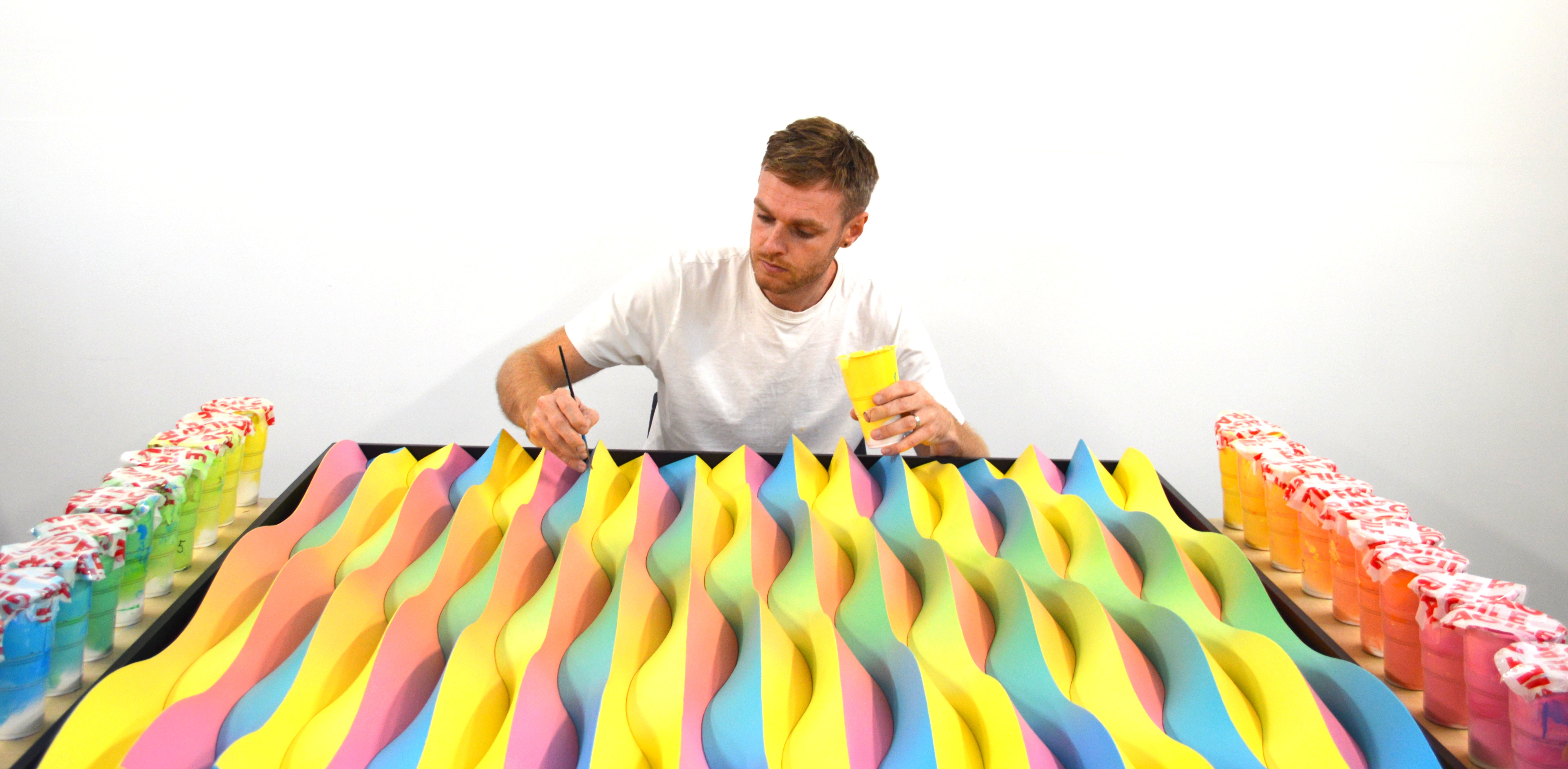 Black and white photo of artist Jack Tanner working on a striped, geometric artwork at a table, with two of his bold, wavy-patterned pieces hanging behind him.