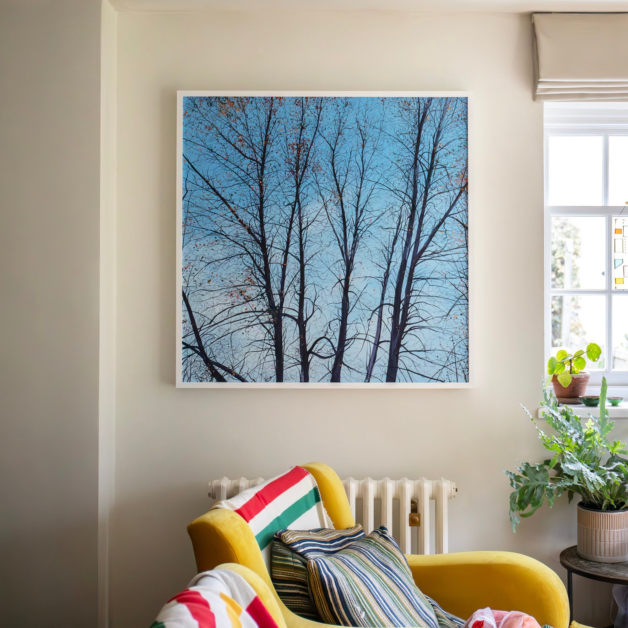 Yellow armchair with colorful cushions in a room with a framed picture of trees on the wall.