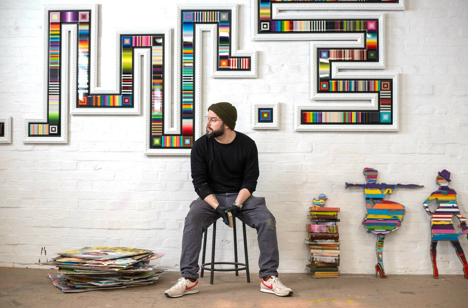 Urban pop artist Lhouette in his studio, seated in front of colourful geometric artwork and mixed media figures, with prints stacked nearby.