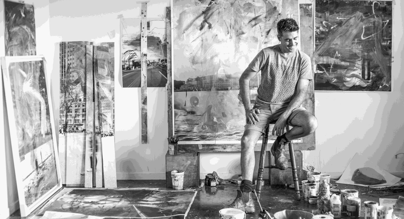 Black-and-white photo of artist Alberto Sanchez sitting in his studio, surrounded by large abstract paintings and paint supplies.