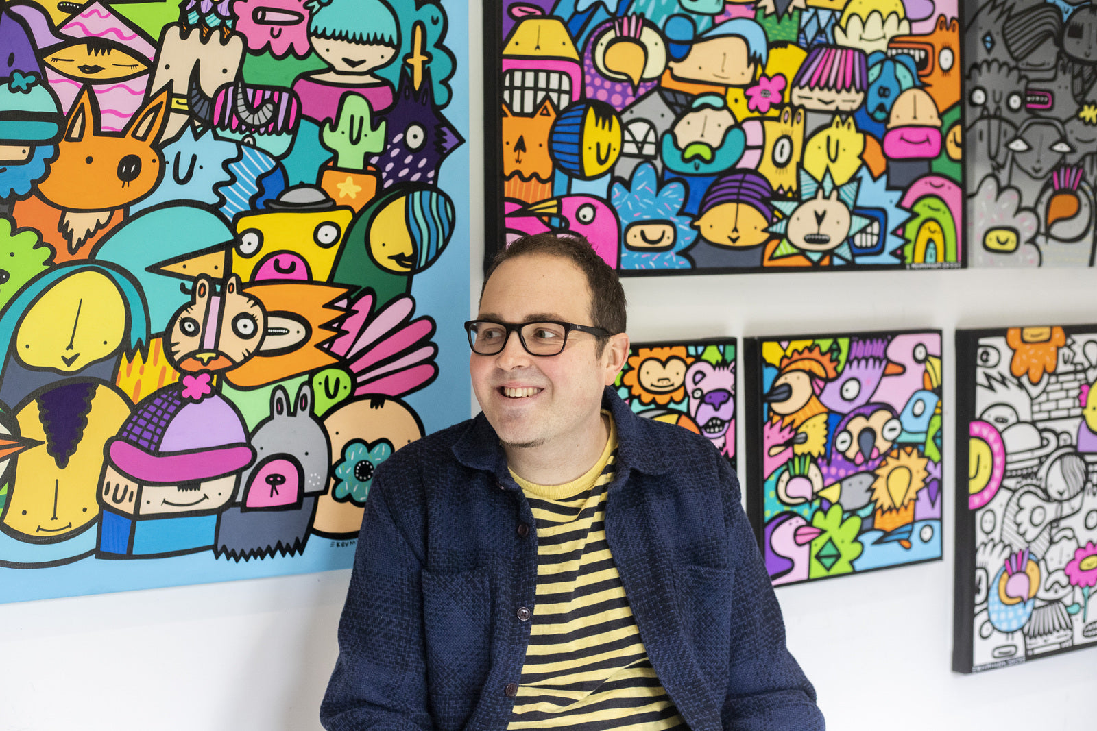 Kev Munday smiling in front of a wall filled with his colourful, character-packed artworks featuring bold outlines and playful faces.