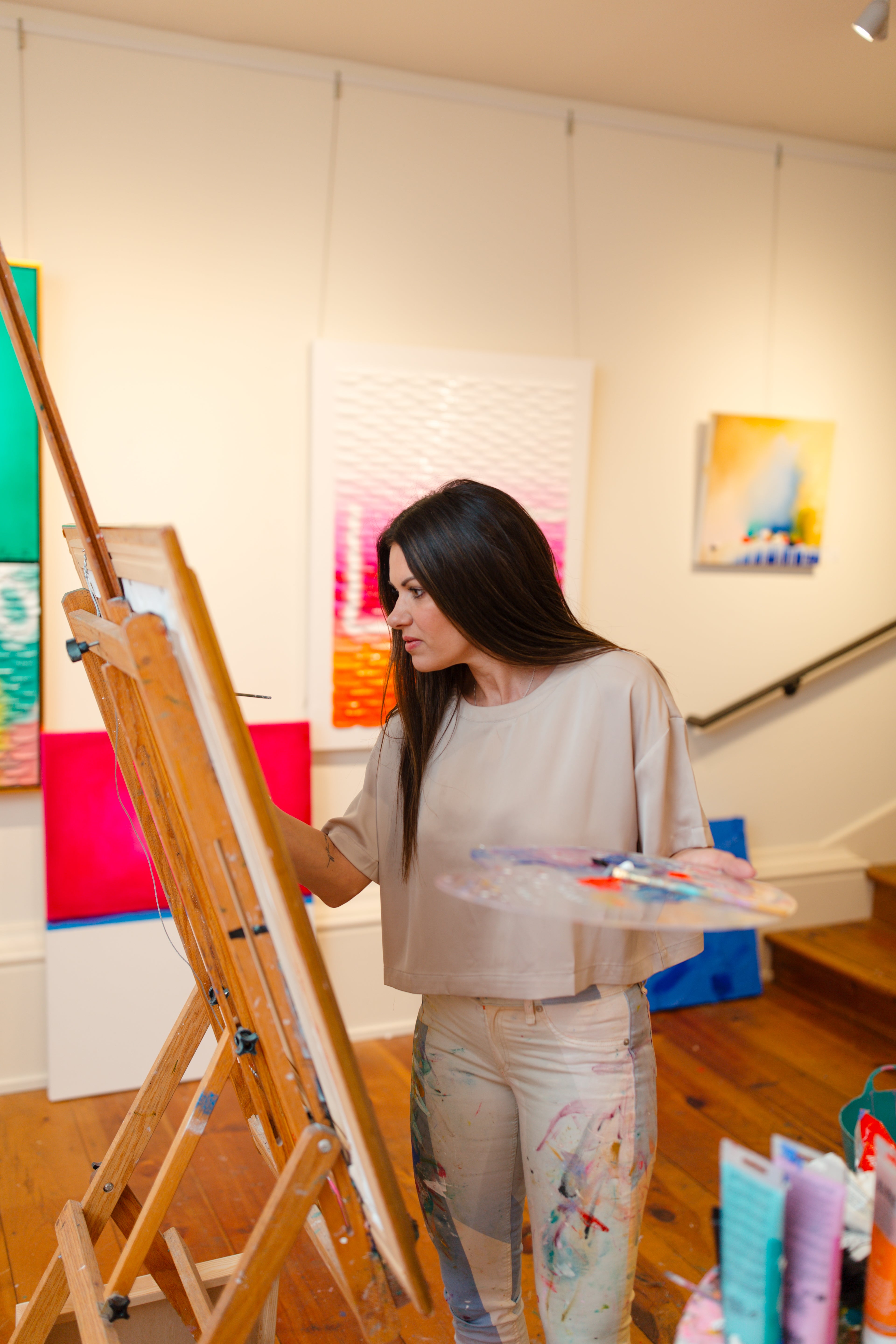 Piper Bridwell painting at an easel in her studio, surrounded by colourful abstract artworks on display